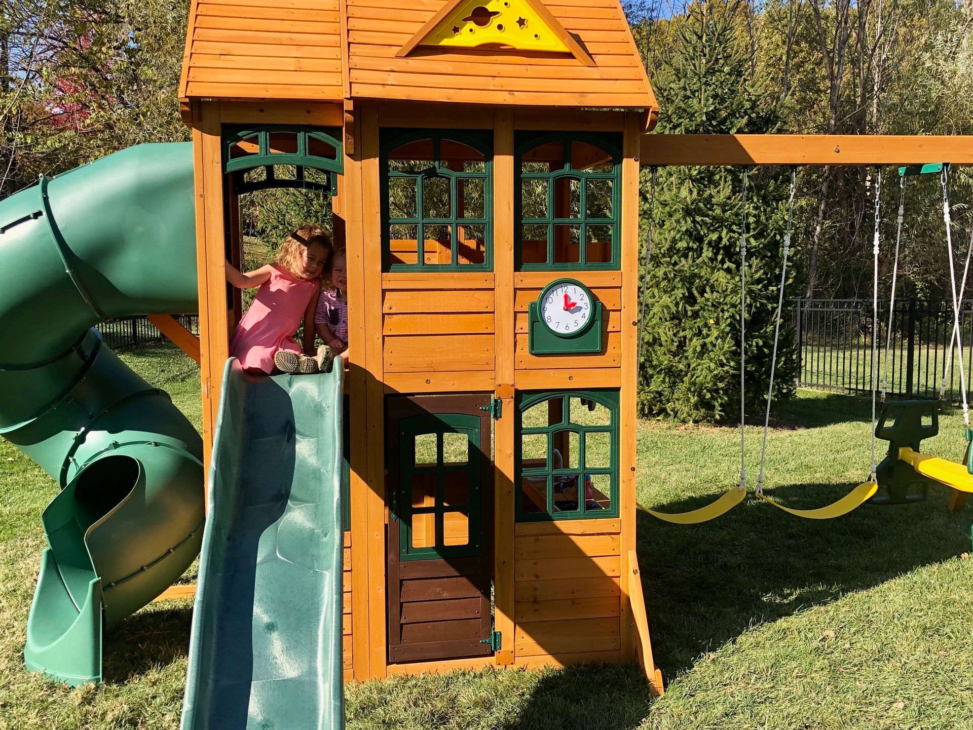 Child sliding down a green slide from a wooden playhouse with a swing set in a sunny backyard.