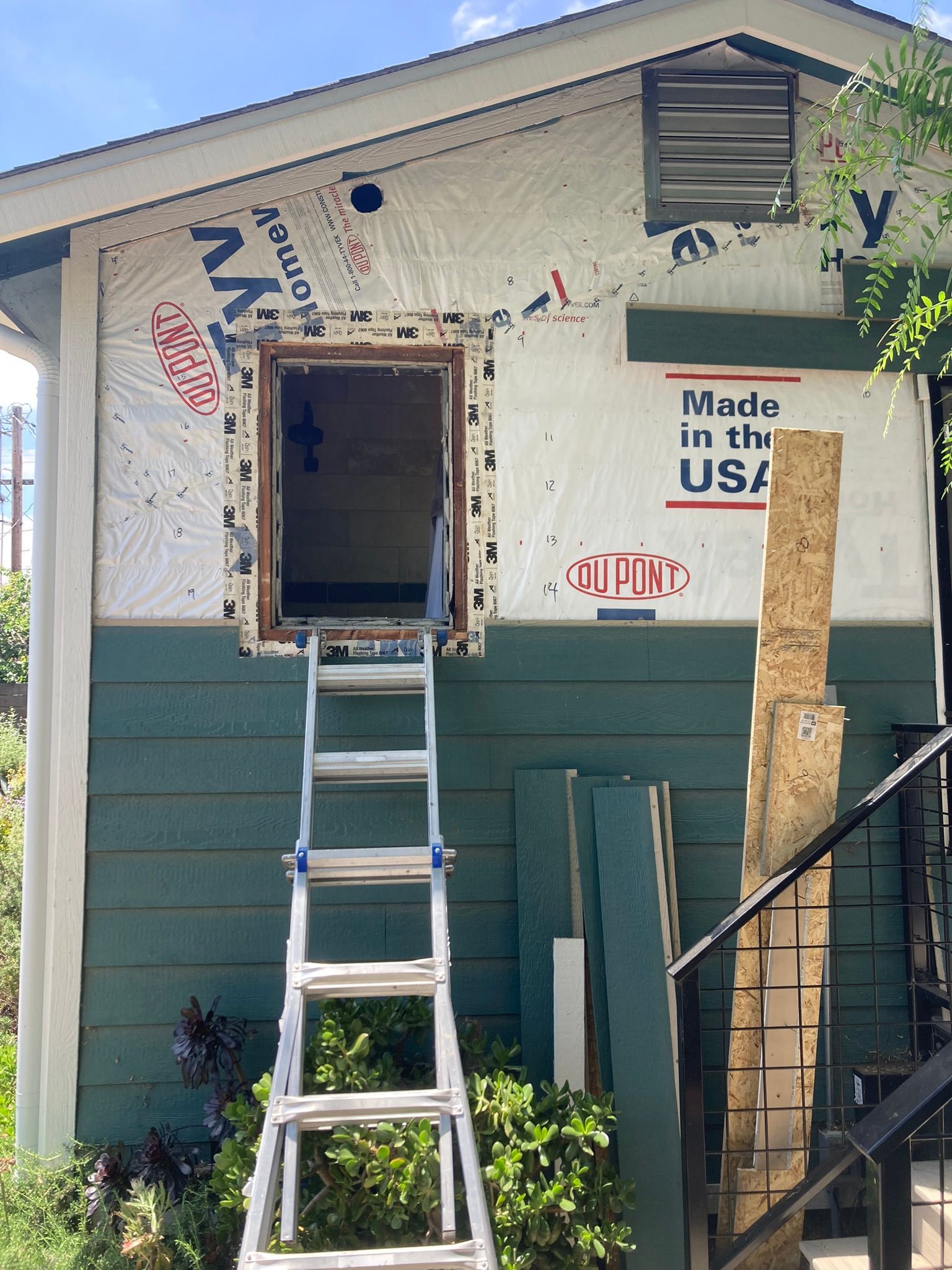 Ladder leading to an open window on a house under construction. Green siding, white wrap, USA sticker.