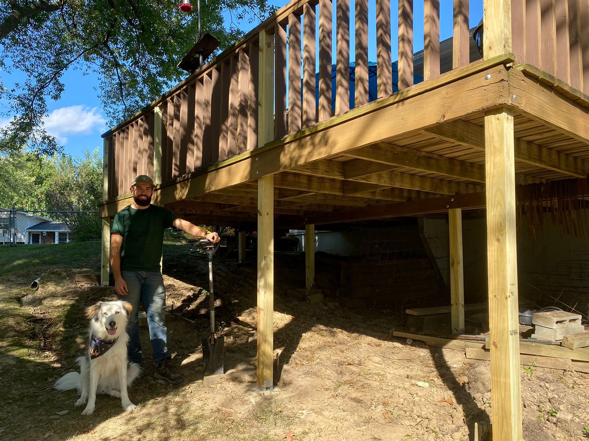 Man and dog stand under a wooden deck. The man smiles, holding a tool. Sunny outdoor setting.