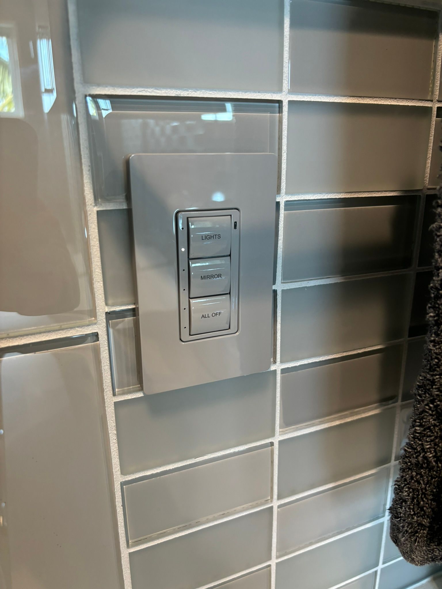 Gray light switch on a tiled wall. Three buttons labeled 