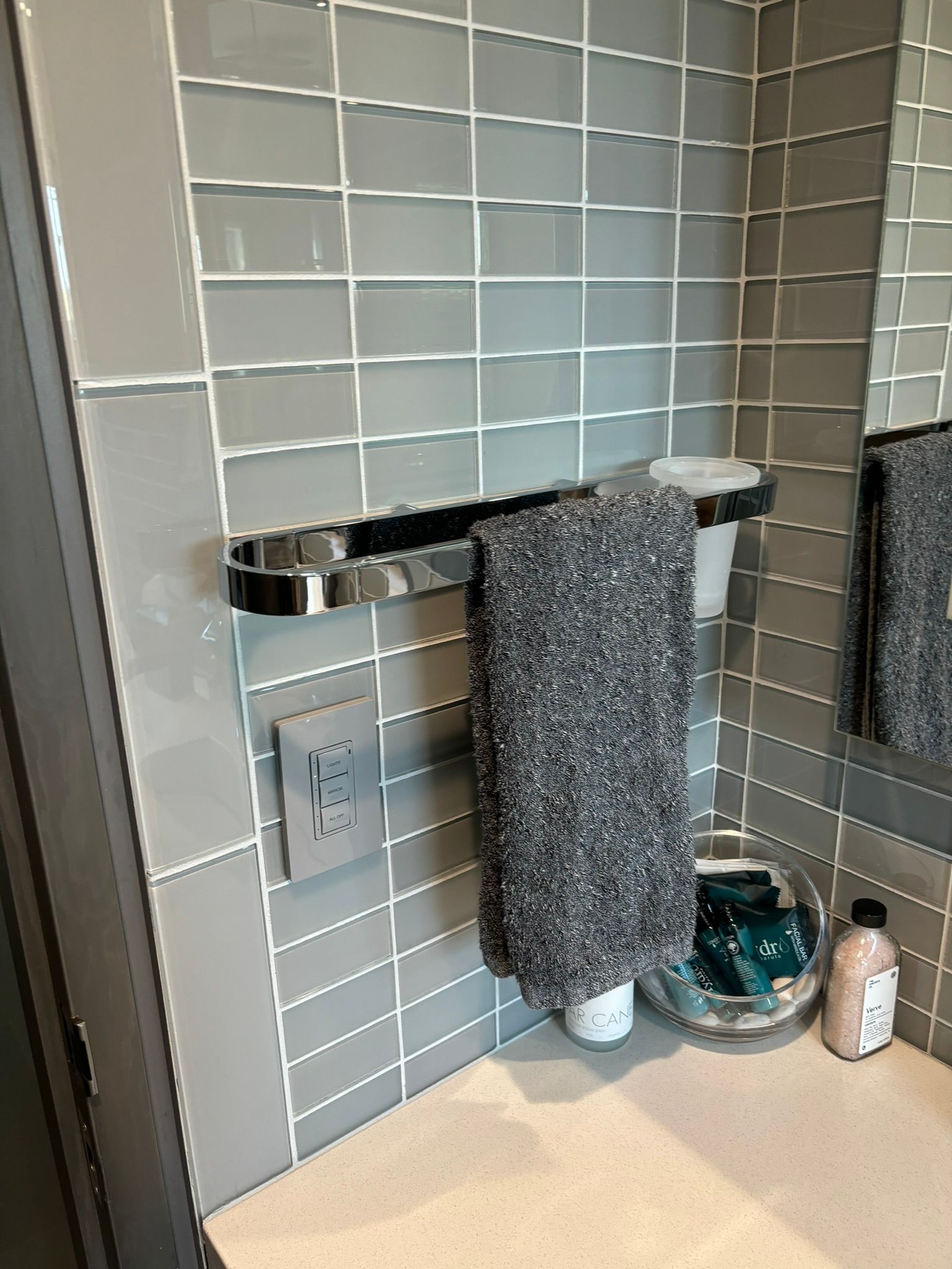 Gray tiled bathroom wall with towel rack, towel, and toiletries.