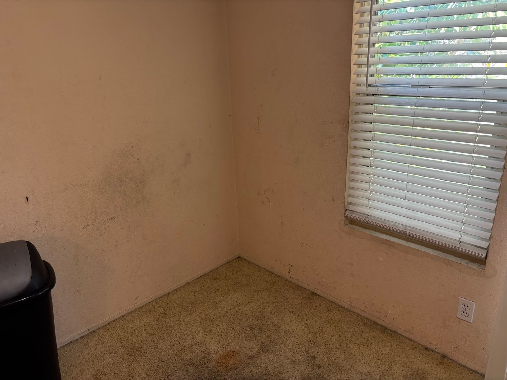 Small room with pink walls, dirty speckled floor, and a window with blinds. Black trash can in the corner.
