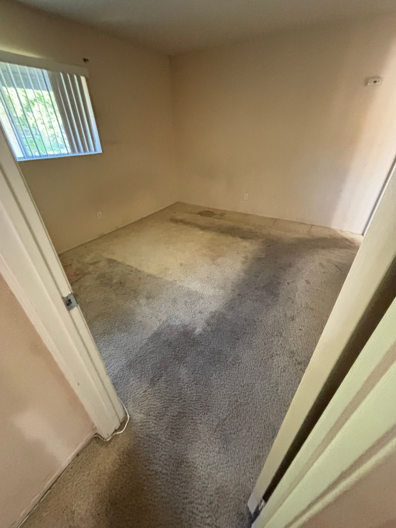 Empty room with stained carpet and small window. Beige walls and doorway.