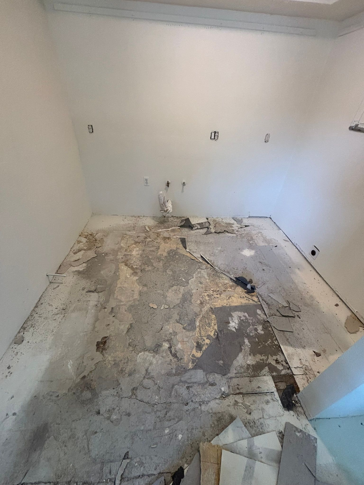 Empty room under construction; damaged floor, white walls, exposed electrical outlets.