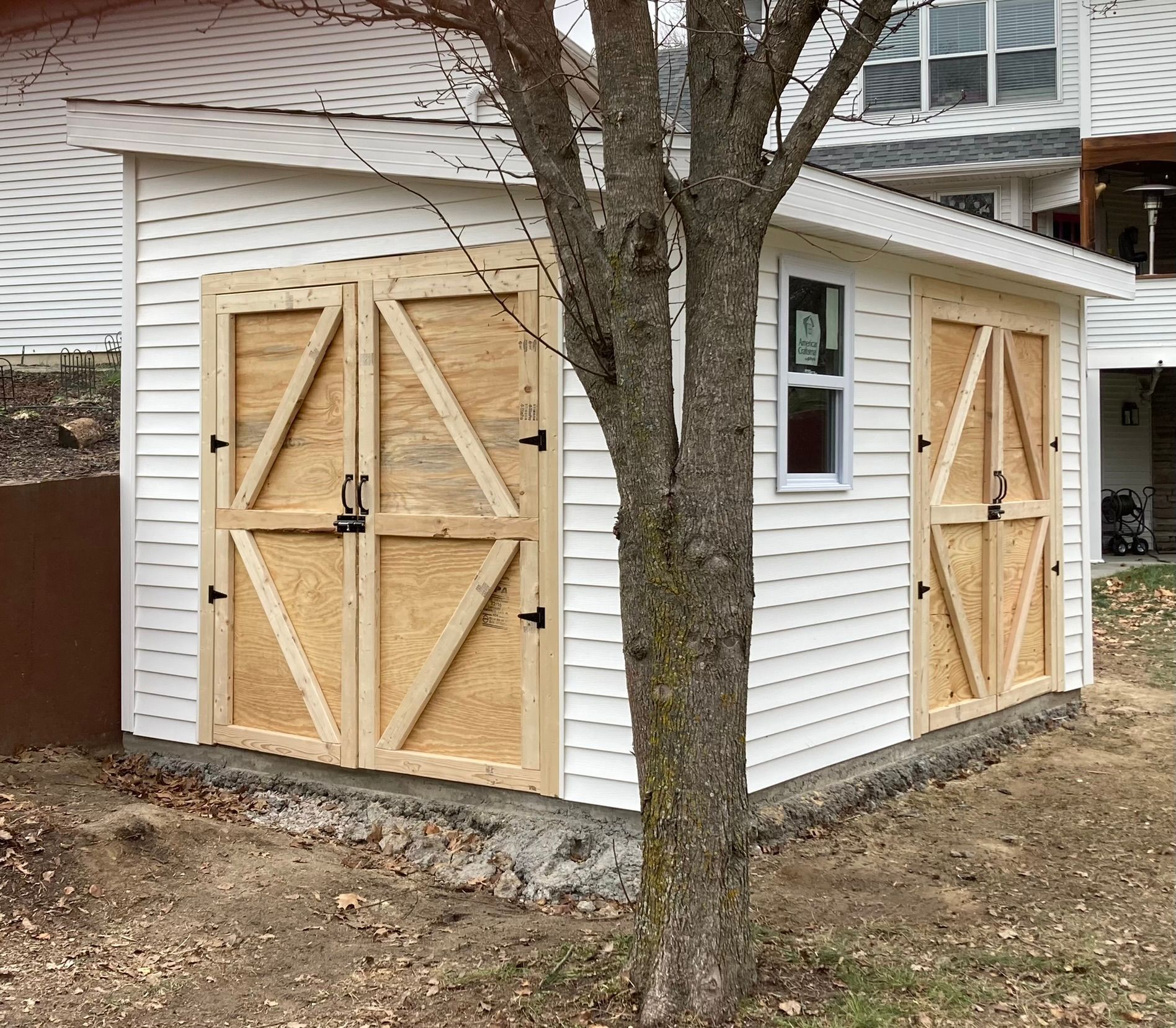 A white shed with wooden doors is next to a tree