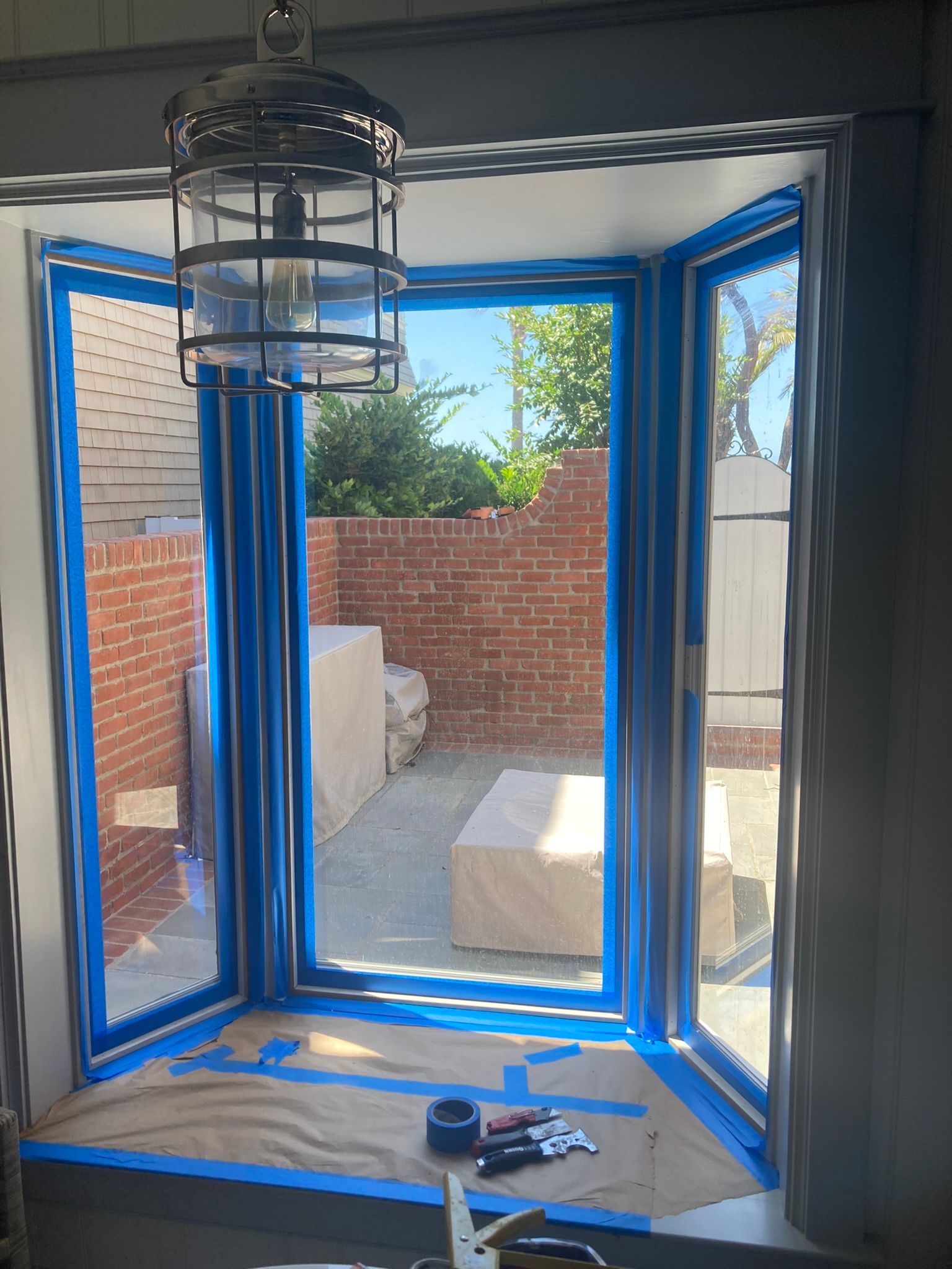 Bay window prepped for painting with blue painter's tape; a brick patio is visible outside.
