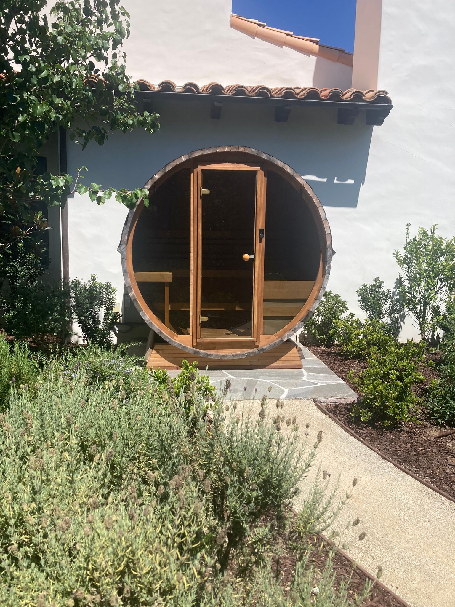 Wooden barrel sauna with open door, set amongst greenery near a building.