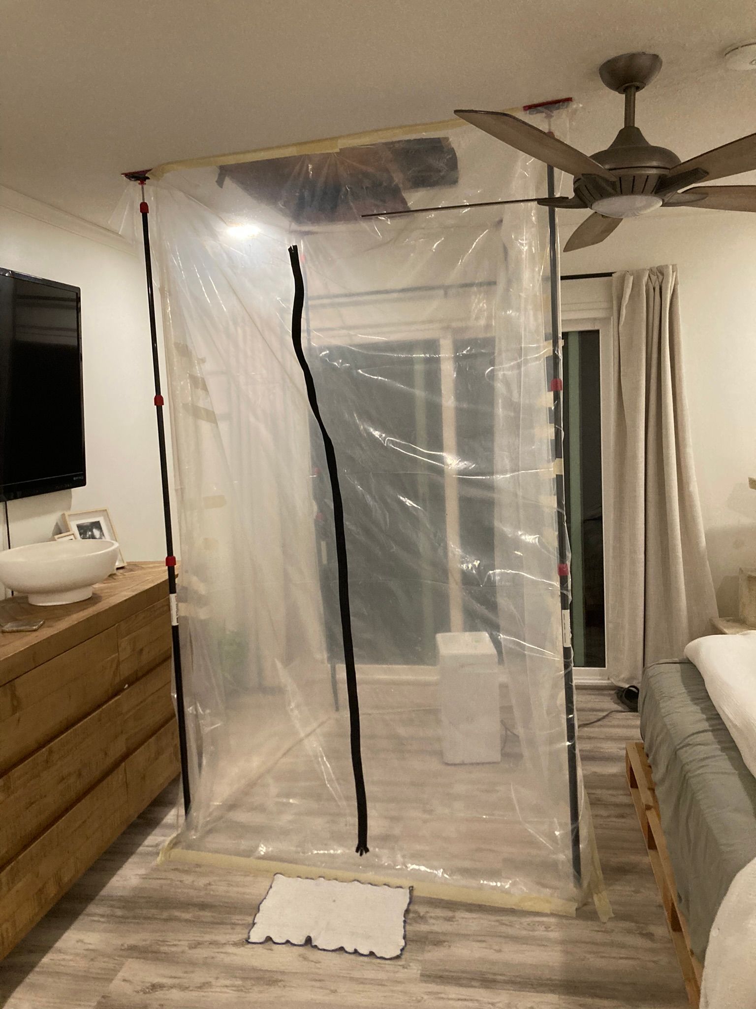 Clear plastic barrier installed in a bedroom, possibly for construction or isolation. Wooden frame, floorboards, and ceiling fan are also visible.