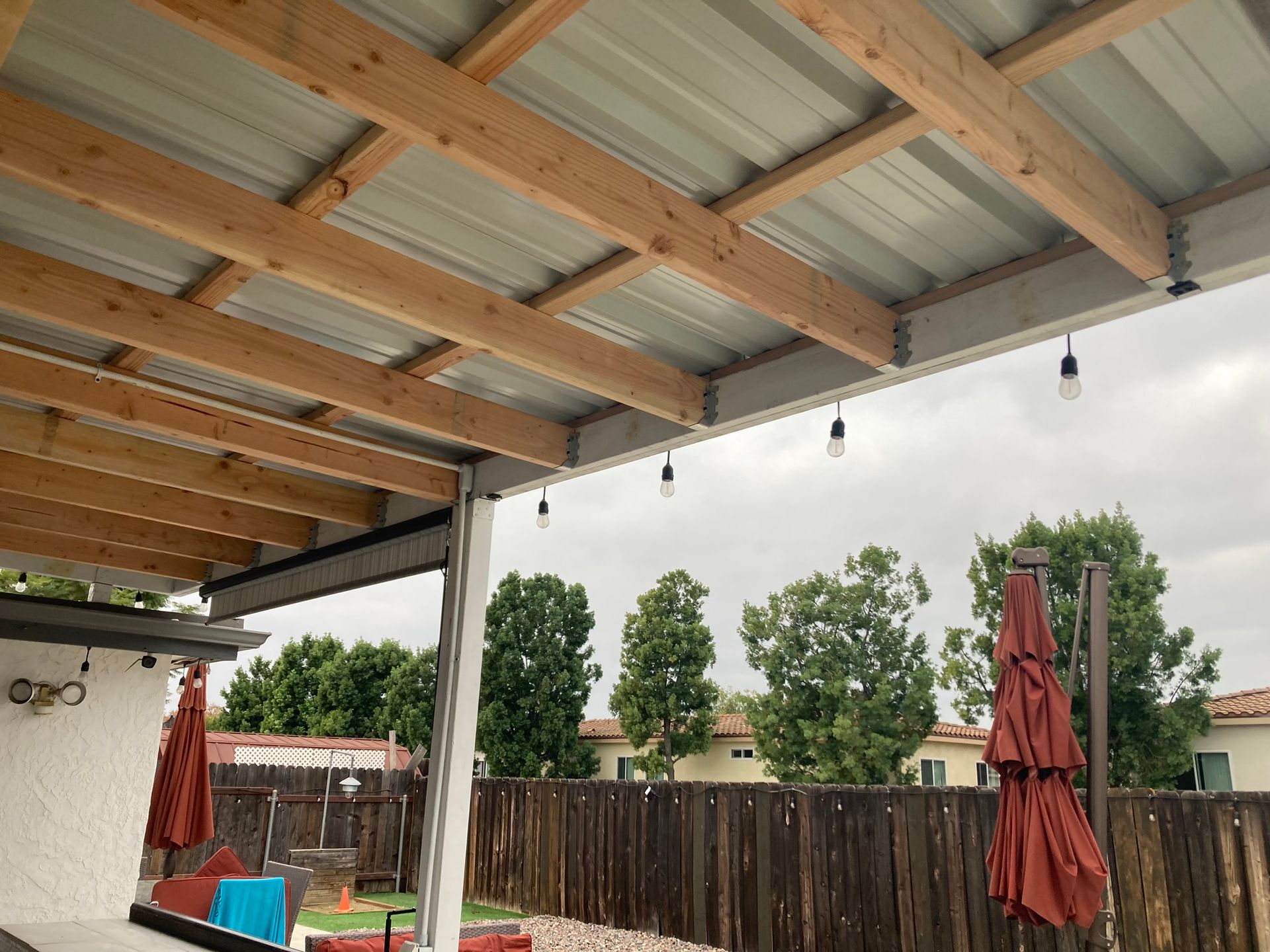 Wooden patio cover with metal roof and string lights, overlooking a backyard with trees and a fence.
