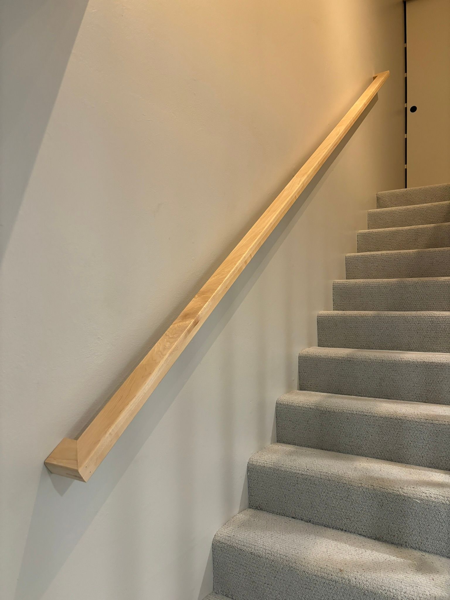 Wooden handrail along a staircase with gray carpeted steps and a light gray wall.