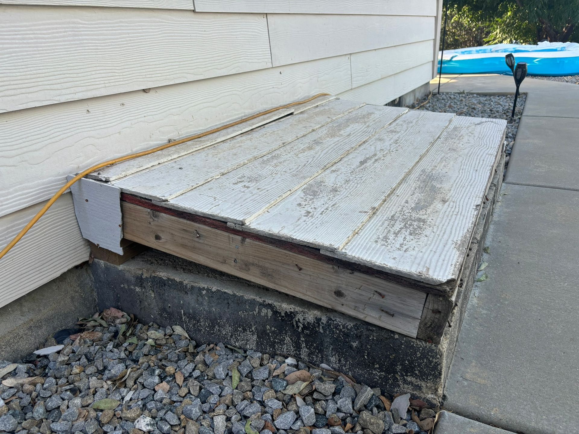 Wooden hatch over a concrete foundation next to a house wall. Gravel and a walkway are visible.