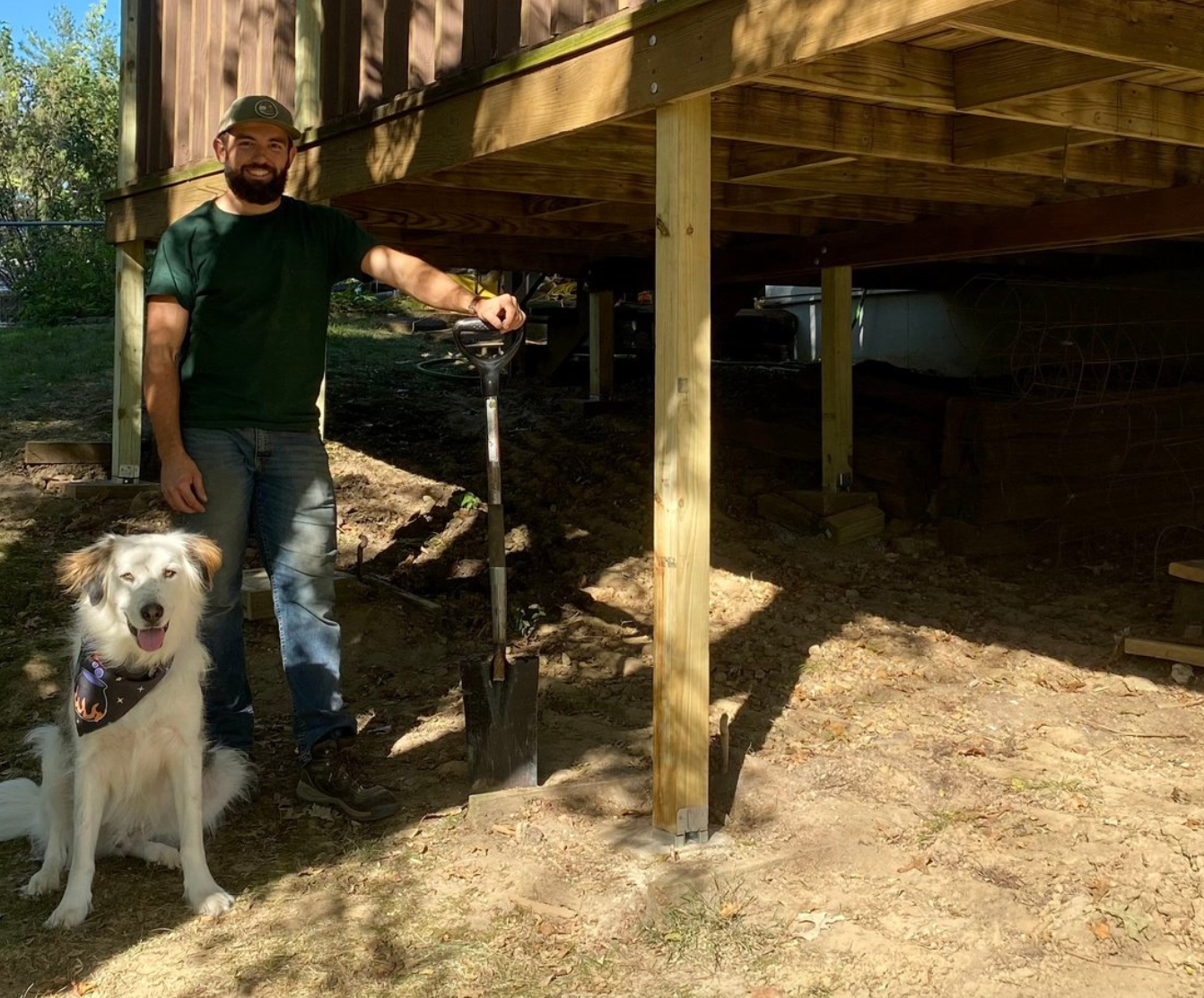 Man and dog under a deck with a shovel. The man wears green, blue jeans; the dog has a bandana.