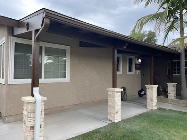 Beige house with brown trim and stone columns, covered patio, and green lawn.