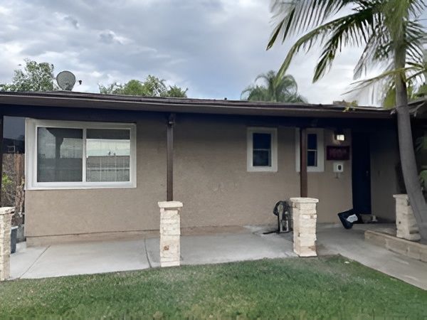 Tan house with a porch, supported by stone columns, under a brown roof. A lawn and driveway in front.
