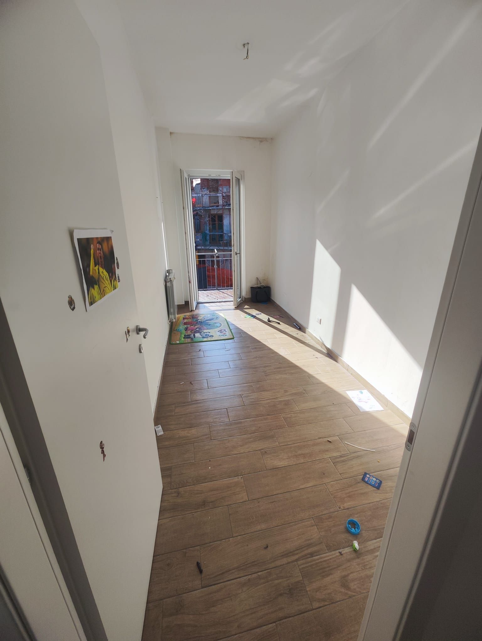 Stretto corridoio bianco con pavimento in legno, porta aperta e luce del sole che filtra da una porta in lontananza.