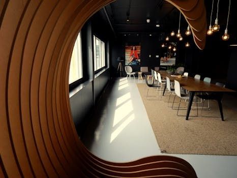 Wooden wave-like wall frames a modern office space with a long table, chairs, and windows with sunlight.