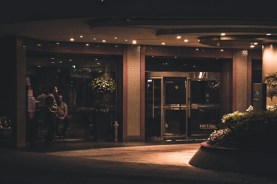 Hotel entrance at night, glass doors, illuminated interior, people visible, dark surroundings.