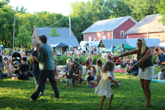 People dance in a sunny, grassy park during an outdoor community event with red barns in the background.
