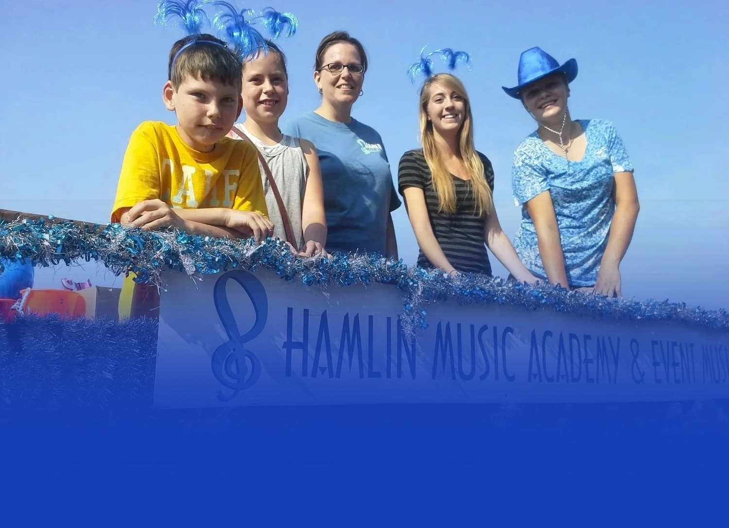 Five people stand on a parade float decorated with blue garland and a banner reading Hamlin Music Academy & Event Center.