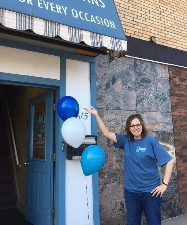 A person wearing a blue t-shirt gestures toward blue and white balloons outside a building entrance.