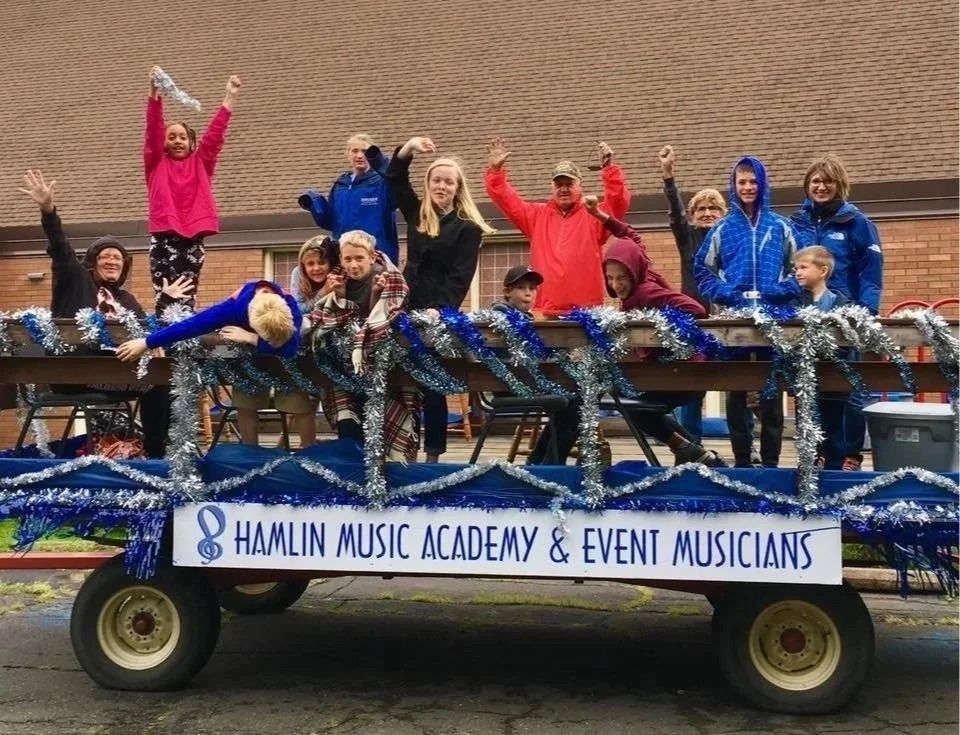 Group of people on a parade float for Hamlin Music Academy & Event Musicians waving and smiling outdoors.