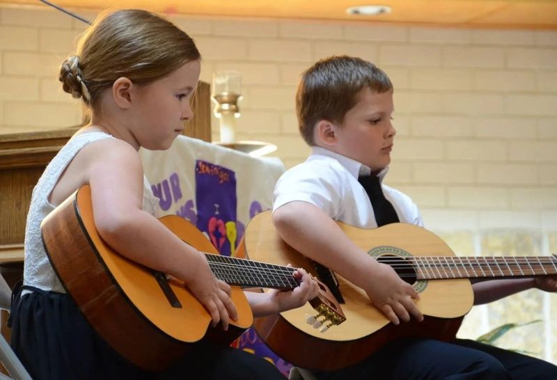Two children sit side-by-side playing acoustic guitars in an indoor setting.