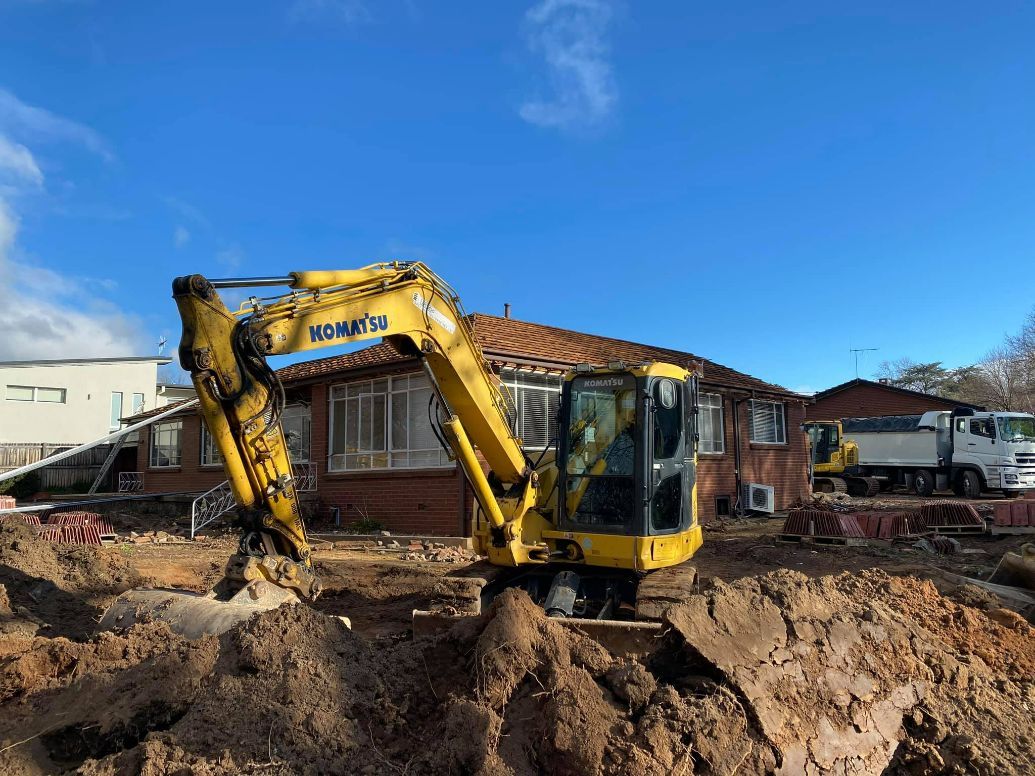 Yellow Komatsu Excavator Digs Earth in Front of a Brick House Under a Blue Sky — Brucic Construction in Dickson, ACT