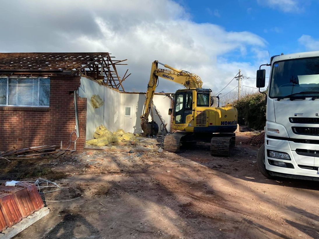 a Yellow Excavator Demolishes a Building Next to a White Truck — Brucic Construction in Dickson, ACT