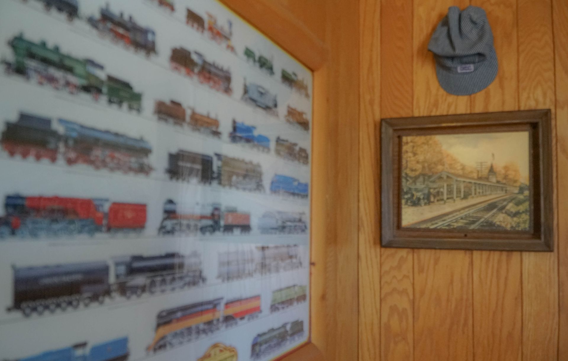 A painting of trains is hanging on a wooden wall next to a hat.