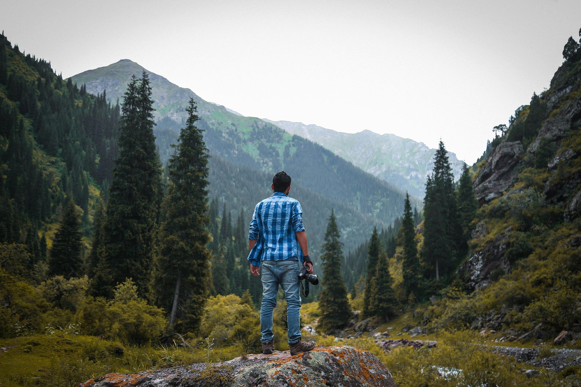 A man in a plaid shirt is standing on a rock in the mountains.