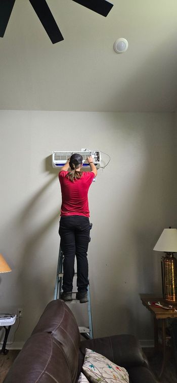 A woman is standing on a ladder fixing an air conditioner in a living room.