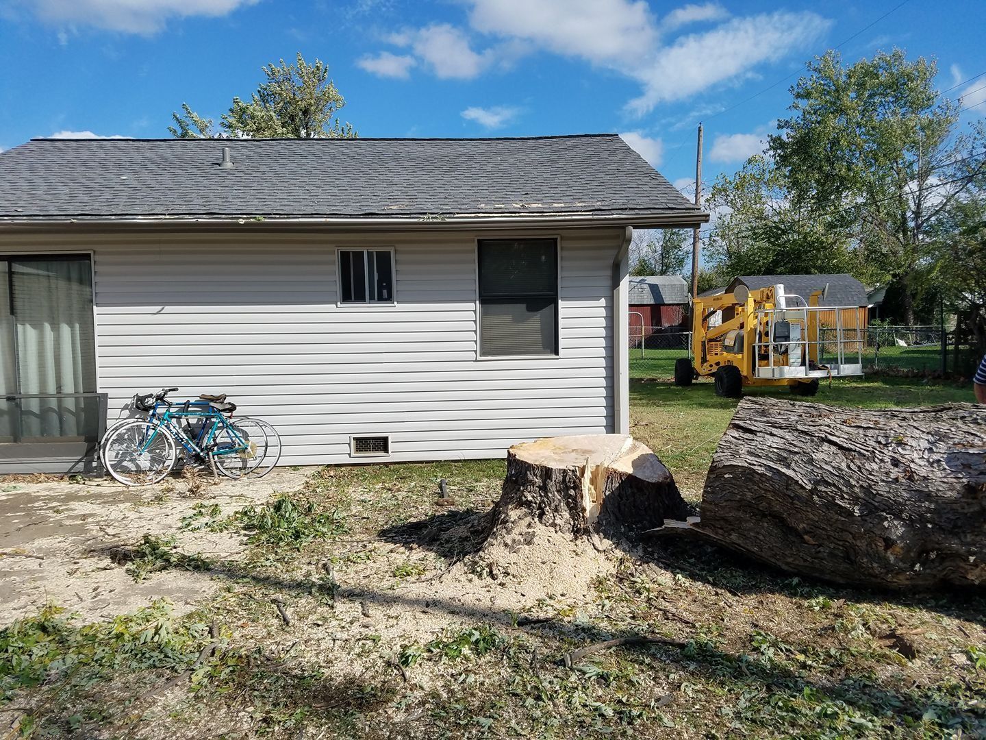Tree stump and log in front of a small building; a wood chipper is visible in the background.