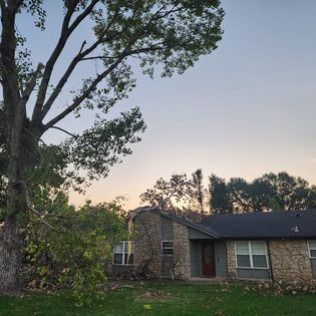 A house with a stone facade sits under a large tree with a cloudy sky.