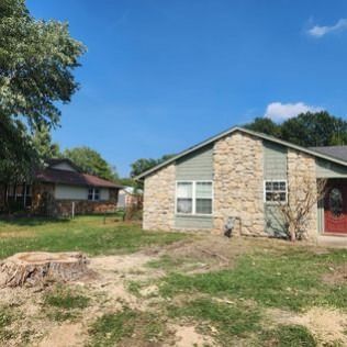 A house with stone facade and a red door sits under a blue sky, a tree stump in the foreground.