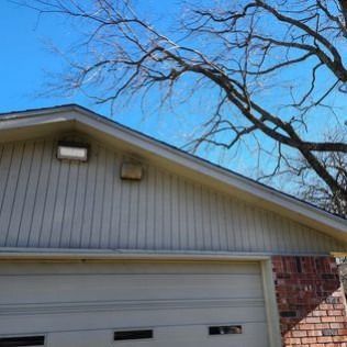 Garage with tan siding and a brick pillar, topped by a roof. Bare tree branches reach across a blue sky.