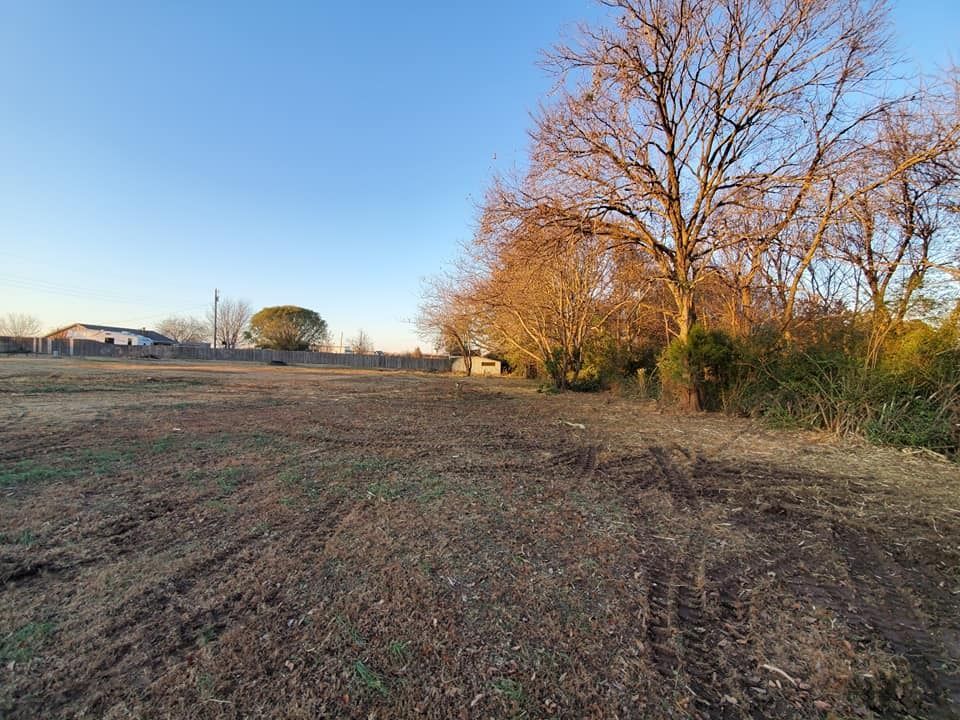 Open field with dirt, trees, and buildings under a blue sky.