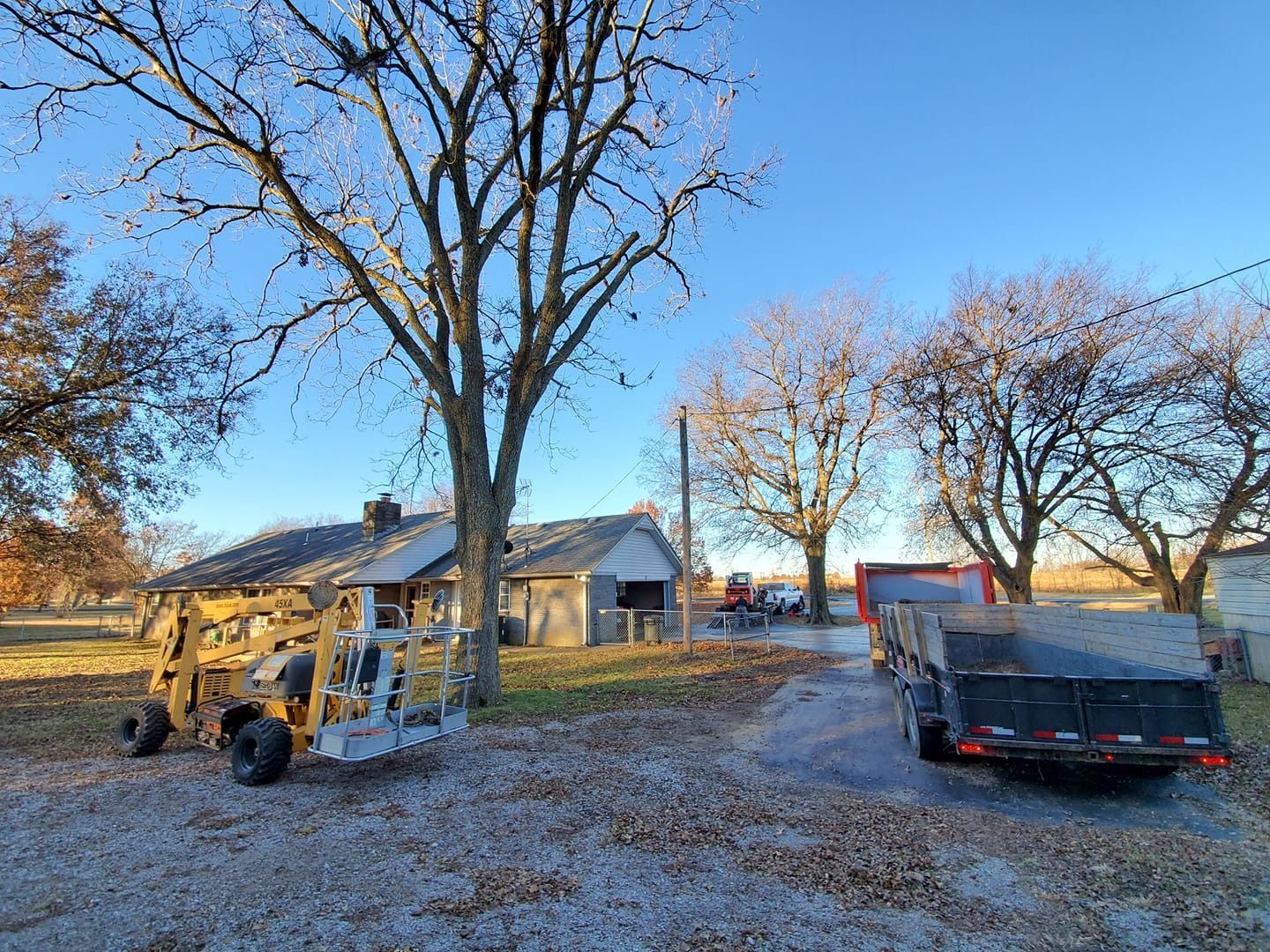 Farm scene with house, trees, and equipment under a clear sky. A trailer and construction vehicle are in the yard.