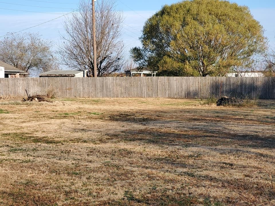 A backyard with dry grass, wooden fence, and trees under a clear blue sky.