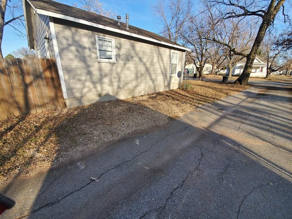 Gray building with a white door and window sits next to a cracked asphalt driveway and brown fence.