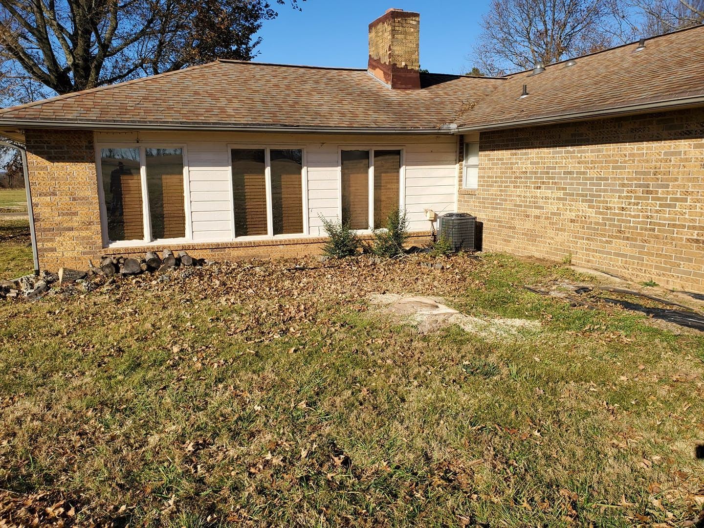 Tan brick house with windows, chimney, and overgrown yard covered in leaves.