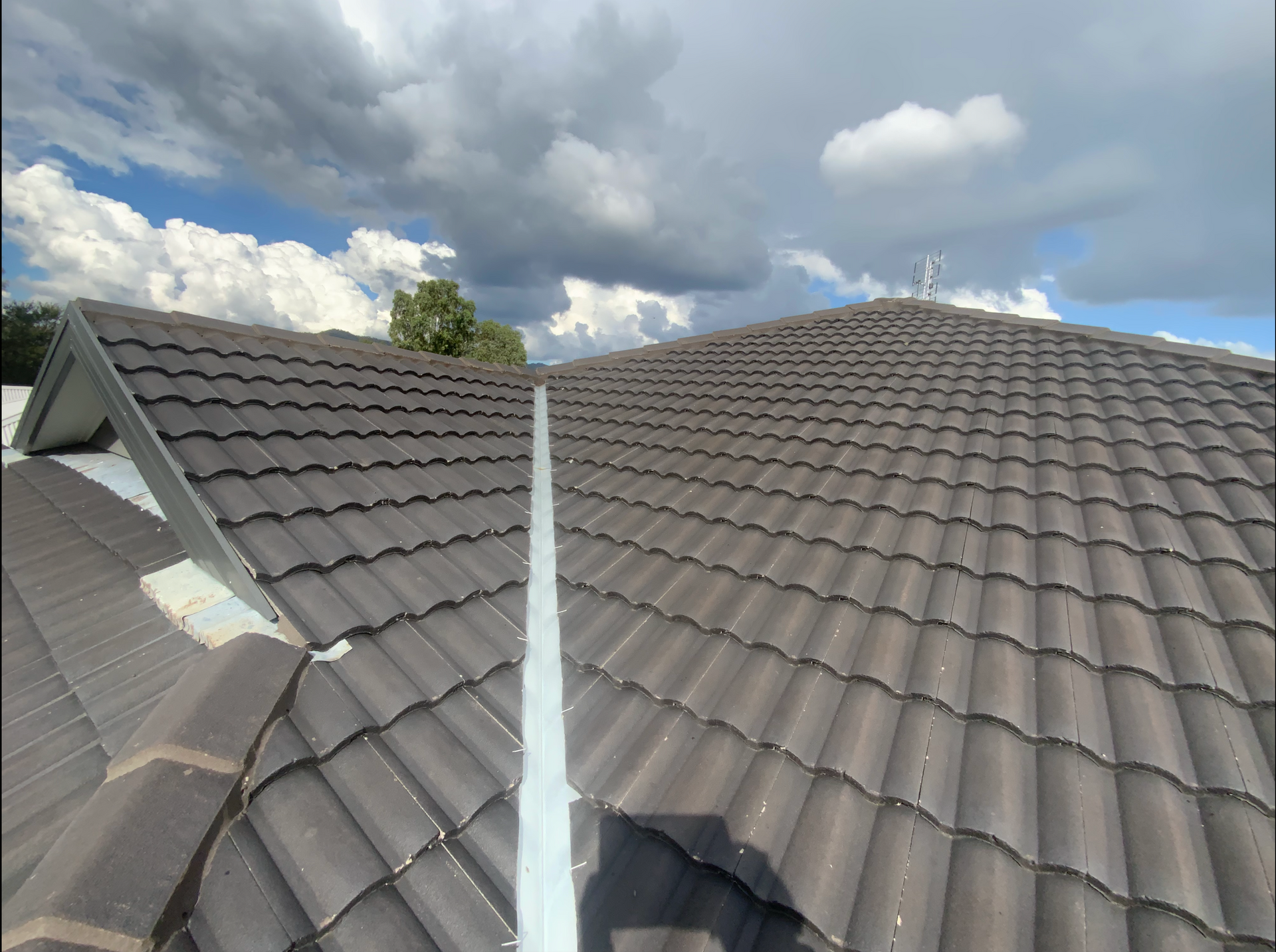 Close-up view of a clean, dark tiled roof under partly cloudy skies. A central metal valley flashing runs down the middle of the roofline, with no visible damage or debris. The image suggests a recently inspected or maintained roofing system, suitable for illustrating building or pest inspection services.— Able Building and Pest Inspections In Coffs Harbour, NSW