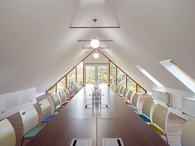 Bright attic meeting room with long table, chairs, skylights, and large windows at one end