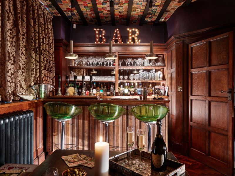 Cozy wood-paneled bar with shelves of bottles, green stools, and a glowing “BAR” sign