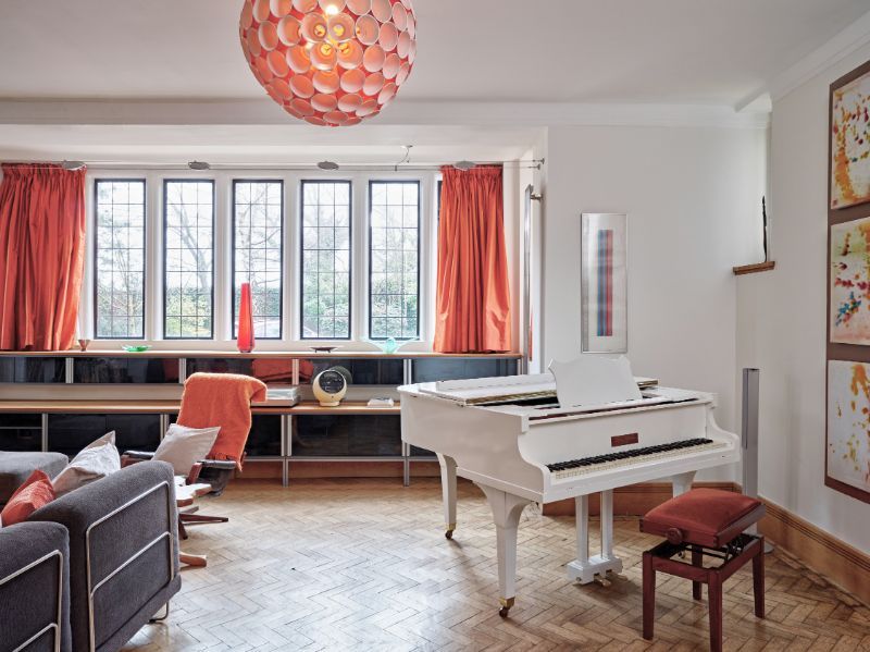 Bright living room with a white grand piano, orange accents, sofa, and large window seat