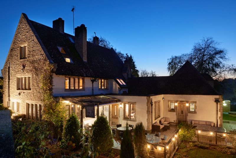 Stone house at dusk with lit windows, warm garden lights, and a blue evening sky