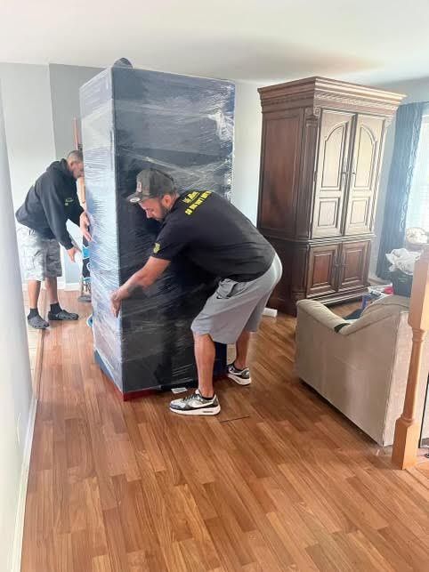 Two movers carrying a large, wrapped cabinet across a room with wood flooring.