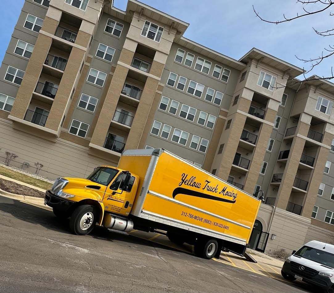 Yellow moving truck parked on a street in front of a multi-story apartment building.