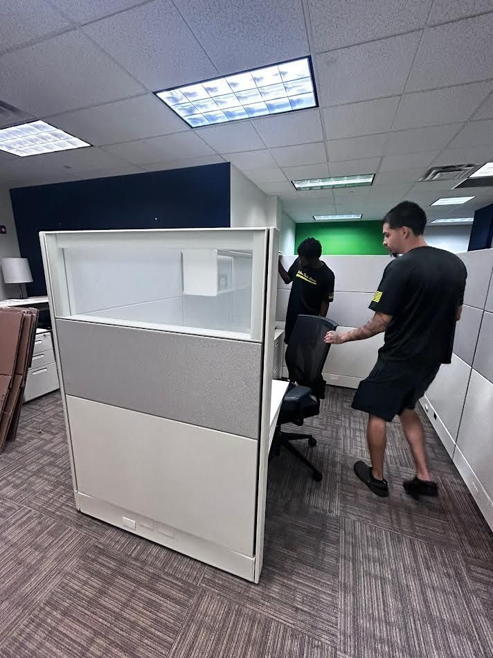 Two people moving office cubicle, in room with carpet and overhead lights.