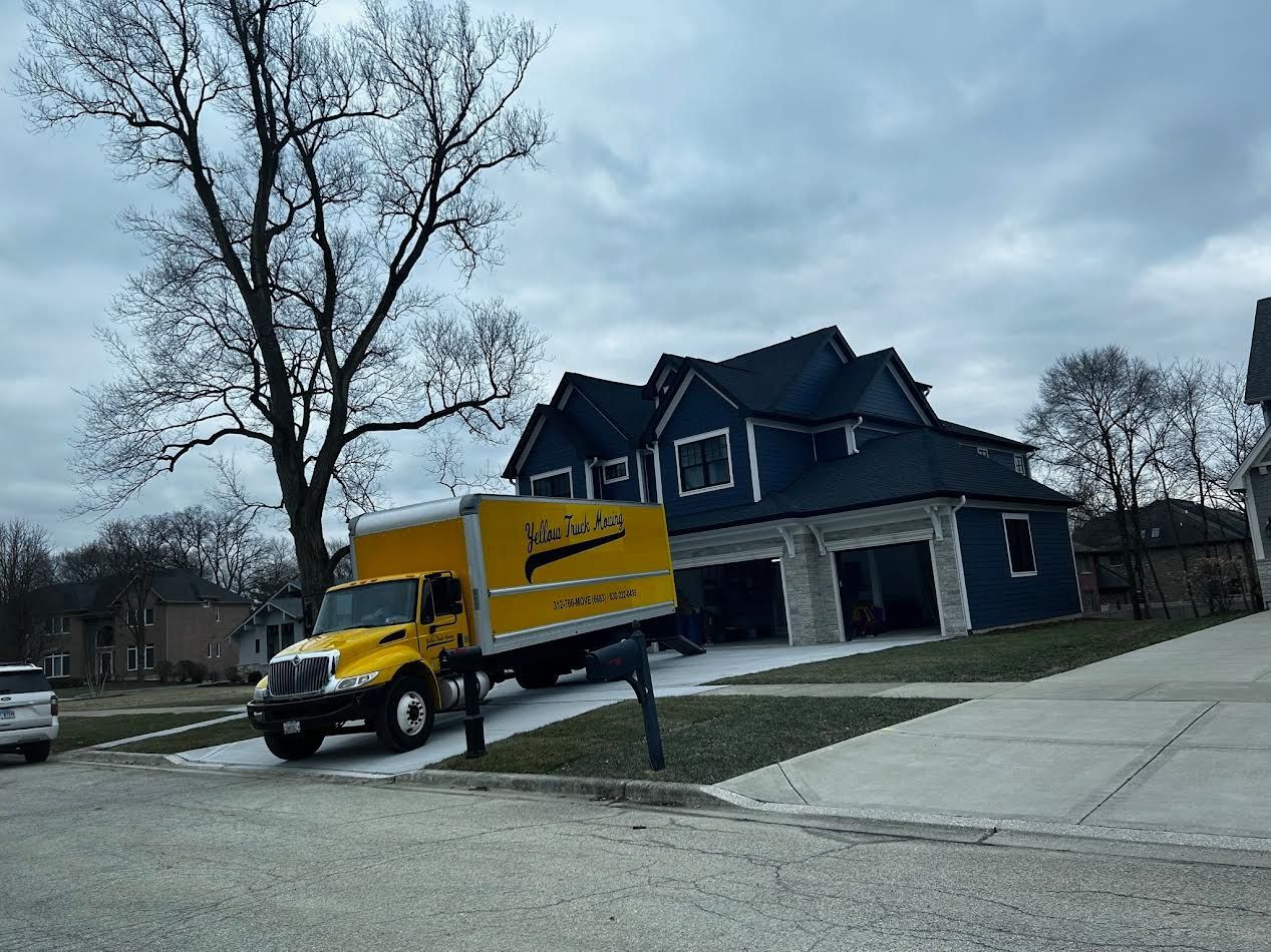 Yellow moving truck parked in front of a blue house on a cloudy day.