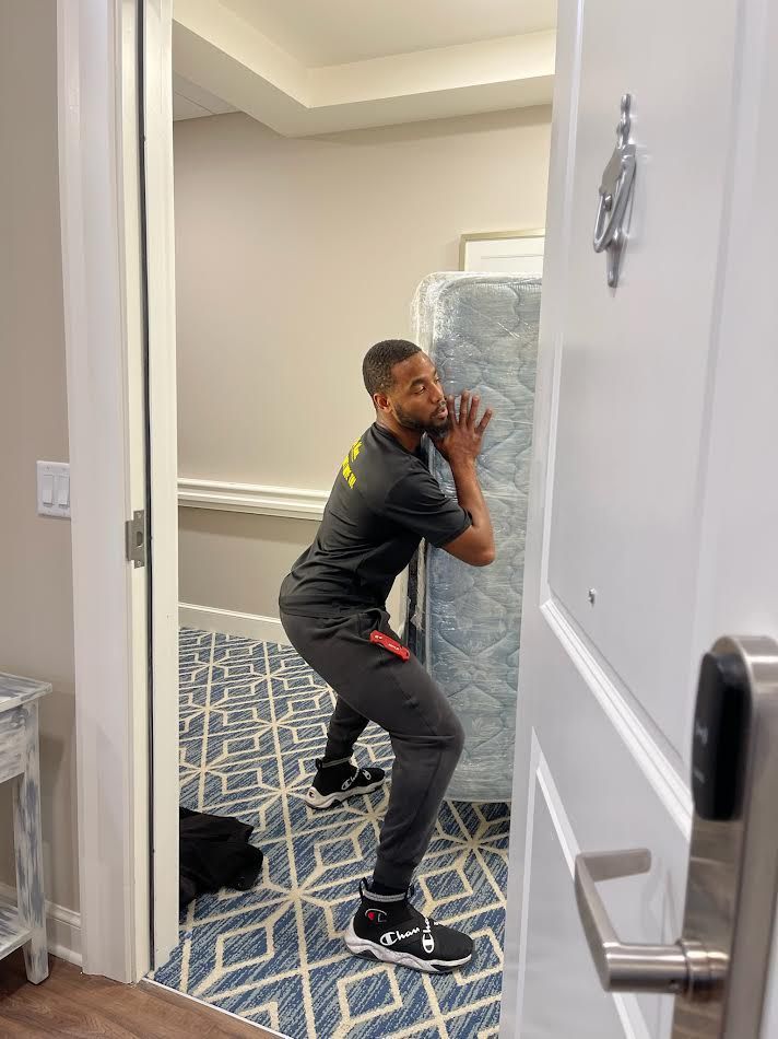 Man struggles to maneuver a mattress through a doorway. He is wearing black athletic wear. The setting is a hotel hallway.